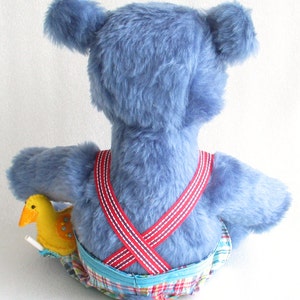 May include: A blue plush toy with a long tail, wearing red and white suspenders and plaid shorts. The toy is holding a yellow plush duck with a red beak.