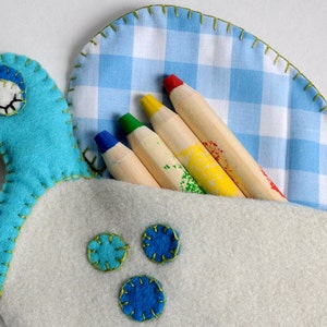 Puede incluir: Un p&aacute;jaro de fieltro azul con un pico amarillo y un ojo blanco, que sostiene un juego de l&aacute;pices de madera de colores en un bolsillo de tela vichy azul y blanco.