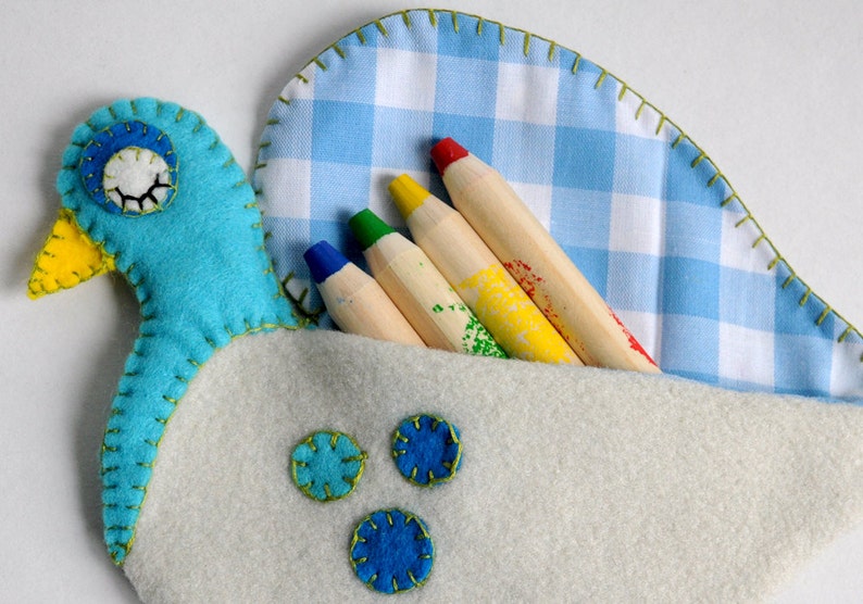Puede incluir: Un p&aacute;jaro de fieltro azul con un pico amarillo y un ojo blanco, que sostiene un juego de l&aacute;pices de madera de colores en un bolsillo de tela vichy azul y blanco.