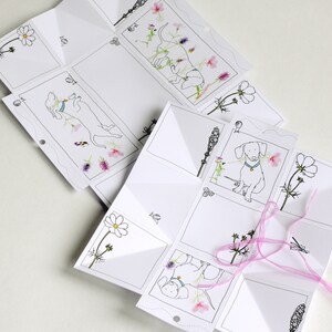 May include: A white paper craft with a pop-up design featuring a dachshund dog and floral illustrations. The paper is folded into a cube shape with a pink ribbon tied around it.