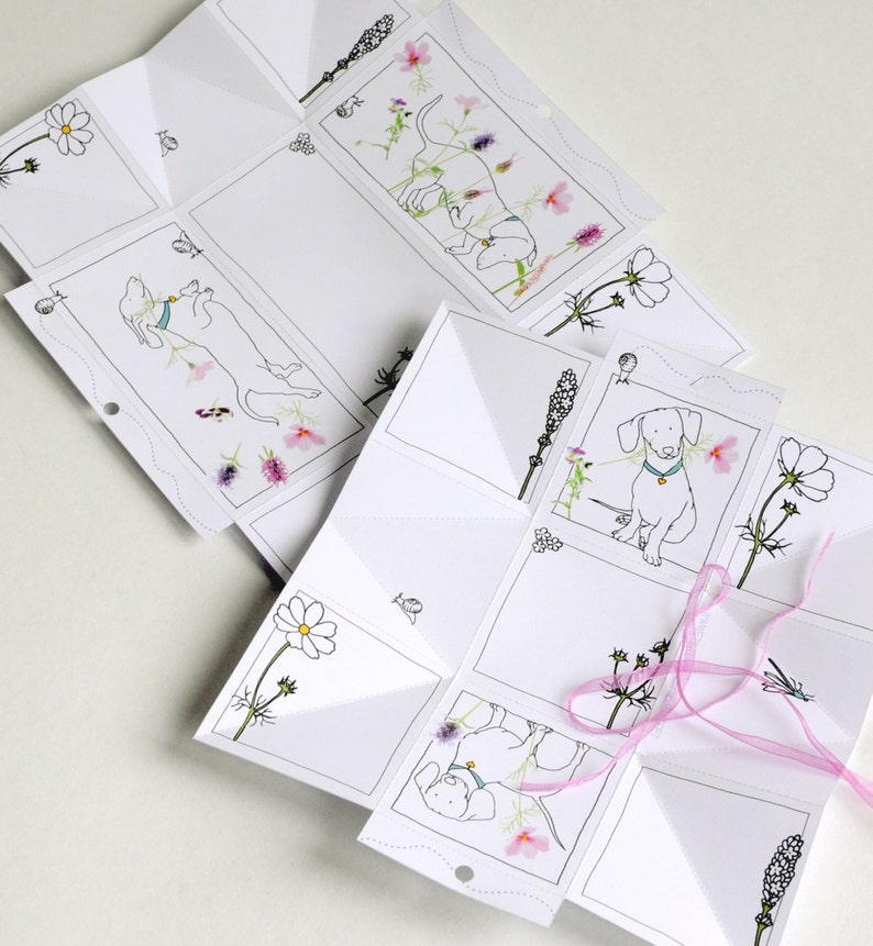 May include: A white paper craft with a pop-up design featuring a dachshund dog and floral illustrations. The paper is folded into a cube shape with a pink ribbon tied around it.