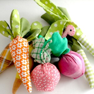 May include: A collection of handmade fabric vegetables, including carrots, onions, and leeks. The carrots have orange and white floral fabric with green polka dot leaves. The onions are pink with white floral patterns. The leeks have green and white checkered stems.