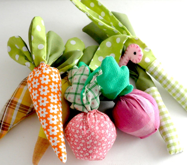 May include: A collection of handmade fabric vegetables, including carrots, onions, and leeks. The carrots have orange and white floral fabric with green polka dot leaves. The onions are pink with white floral patterns. The leeks have green and white checkered stems.