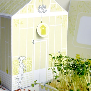 May include: A white cardboard greenhouse with a yellow and green pattern. The greenhouse has a small birdhouse hanging from the roof. There are two owls perched on the roof. The greenhouse is sitting on a table with a pot of green sprouts.