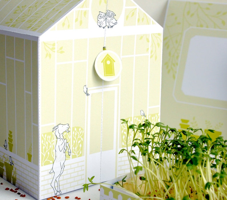 May include: A white cardboard greenhouse with a yellow and green pattern. The greenhouse has a small birdhouse hanging from the roof. There are two owls perched on the roof. The greenhouse is sitting on a table with a pot of green sprouts.