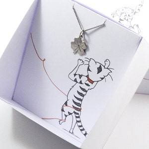 May include: A silver four-leaf clover pendant necklace in a white gift box with a cartoon cat illustration. The box has a red string attached to it.