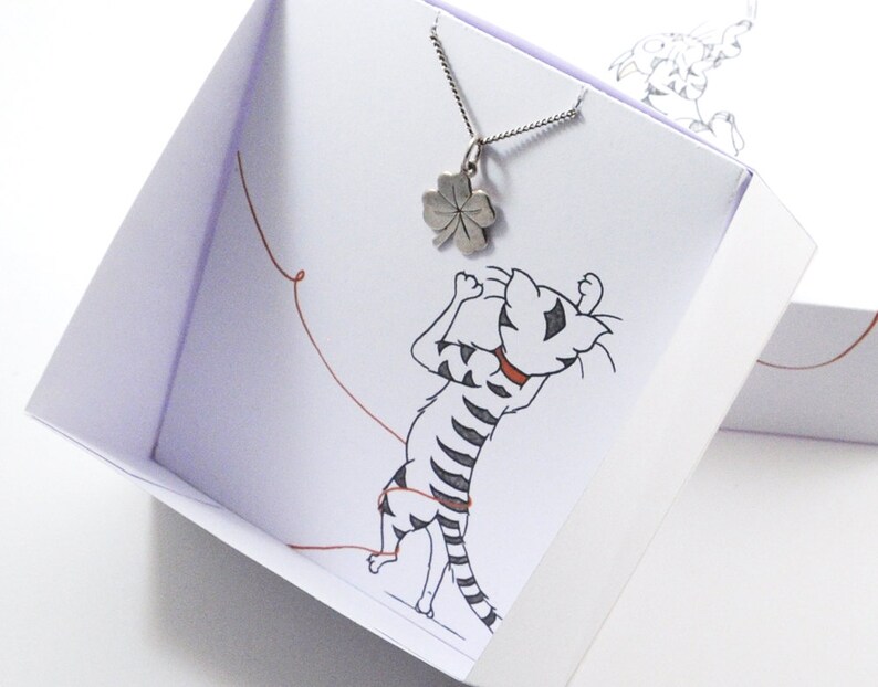 May include: A silver four-leaf clover pendant necklace in a white gift box with a cartoon cat illustration. The box has a red string attached to it.