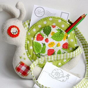 Puede incluir: Un caracol de peluche blanco con una concha de tela verde y blanca. La concha tiene un bolsillo con un forro de tela de cuadros rojos y blancos. El caracol sostiene dos l&aacute;pices de colores y una tarjeta blanca con un dibujo de caracol.