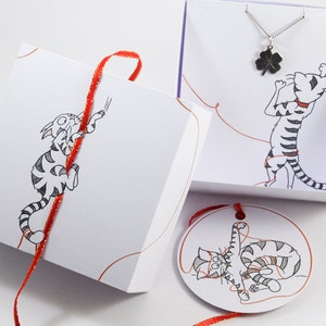 May include: A white gift box with a red ribbon and a black and white illustration of a cat. The box is tied with a red ribbon and has a small tag with a black and white illustration of a cat on it. The tag is attached to the ribbon with a red string.