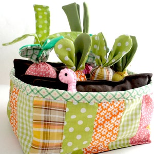 May include: A colorful fabric basket with a patchwork design, filled with plush vegetables. The basket features green, orange, and pink patterned fabrics. The vegetables include radishes and carrots with green fabric tops, and a pink worm.