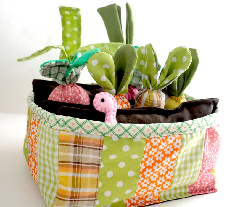 May include: A colorful fabric basket with a patchwork design, filled with plush vegetables. The basket features green, orange, and pink patterned fabrics. The vegetables include radishes and carrots with green fabric tops, and a pink worm.