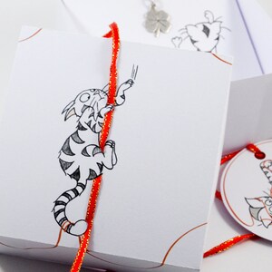 May include: A white gift box with a red ribbon and a black and white illustration of a cat climbing the ribbon. The box is tied with a red ribbon and has a small white tag with a black and white illustration of a cat on it.