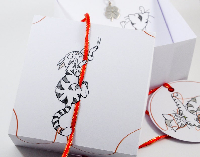 May include: A white gift box with a red ribbon and a black and white illustration of a cat climbing the ribbon. The box is tied with a red ribbon and has a small white tag with a black and white illustration of a cat on it.