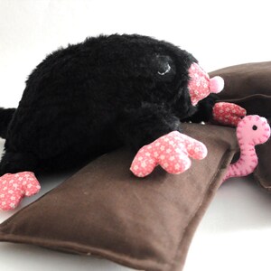 May include: A plush black mole toy with pink floral patterned feet and nose, resting on a brown pillow. A pink worm toy with a stitched face is also present. The toys are designed for children.