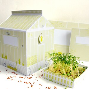 May include: A yellow papercraft greenhouse with a white roof and a small white window. The greenhouse is decorated with green leaves and birds. A small white planter with a white picket fence is in front of the greenhouse. The planter is filled with green sprouts.