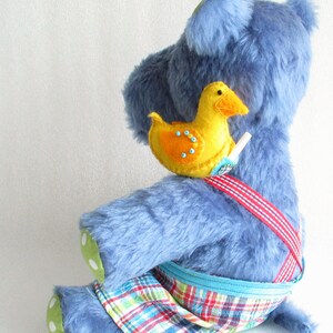 May include: A blue plush toy wearing a red and blue plaid overall with a yellow felt duck attached to the strap. The duck has orange accents and black bead eyes.