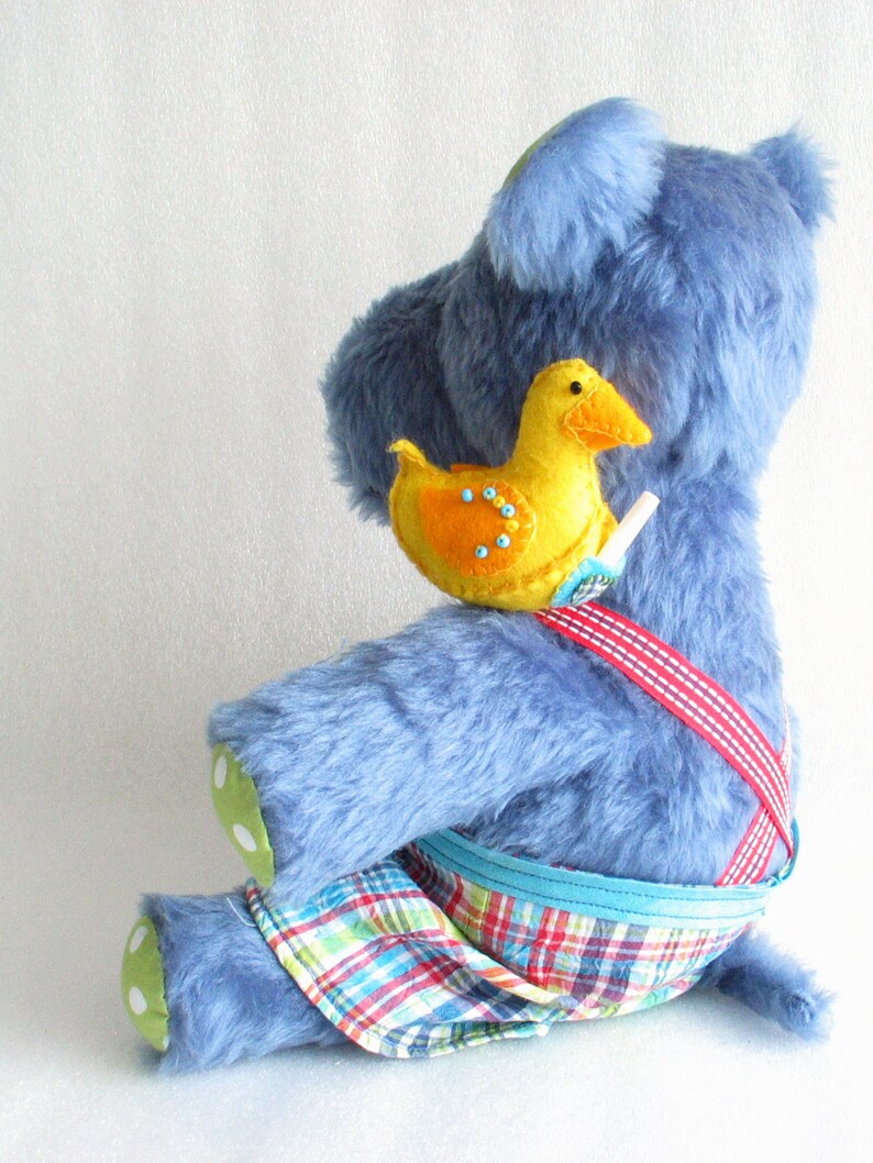 May include: A blue plush toy wearing a red and blue plaid overall with a yellow felt duck attached to the strap. The duck has orange accents and black bead eyes.
