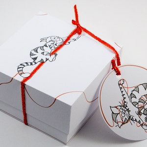 May include: A white gift box with a red ribbon and a cat illustration. The box has a round tag attached with a cat illustration and the text "The Cat's Meow".