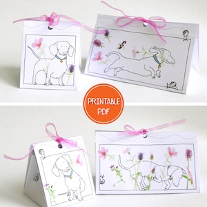 May include: Printable PDF template for a dachshund-themed gift box. The template features a black and white illustration of a dachshund with flowers and a snail. The box is folded into a triangular shape and has a pink ribbon.