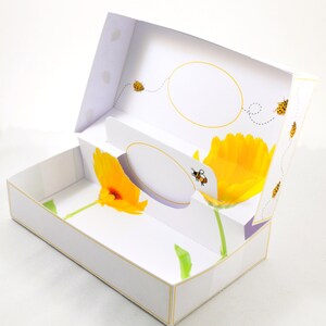 May include: White tissue box with a floral design featuring yellow flowers and a bee. The box has a cutout for tissues and a decorative oval.