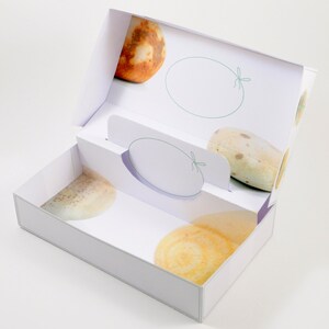 May include: White gift box with a lid open, revealing three round, textured, beige and brown soap bars. The box has a white background with a green ribbon design.