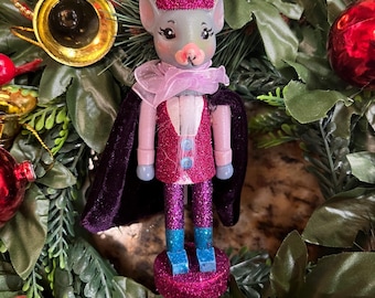 May include: A purple and silver glitter nutcracker with a crown, a pink cape, and blue boots. The nutcracker is a mouse with a pink face and a pink bow tie. The nutcracker is on a green and red wreath.