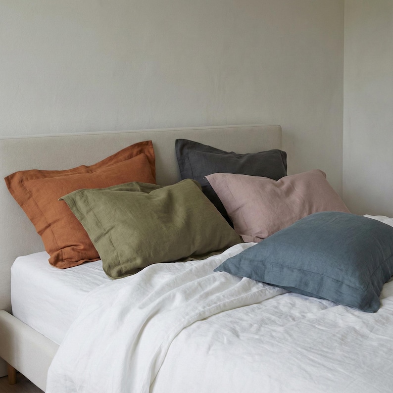 May include: A collection of five rectangular pillow shams in various colors including burnt orange, olive green, charcoal gray, dusty pink, and slate blue. Each pillow sham features a wide flange border and is arranged on a white bed sheet.