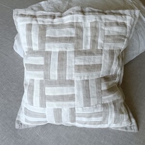 Può includere: Un cuscino quadrato con una fodera in lino bianco e un motivo in lino marrone. Il motivo è una serie di linee orizzontali e verticali che creano un design a griglia.