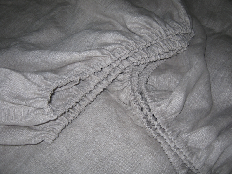 May include: A close-up of a gray fitted sheet with elastic edging. The sheet is made of a soft, natural fabric.