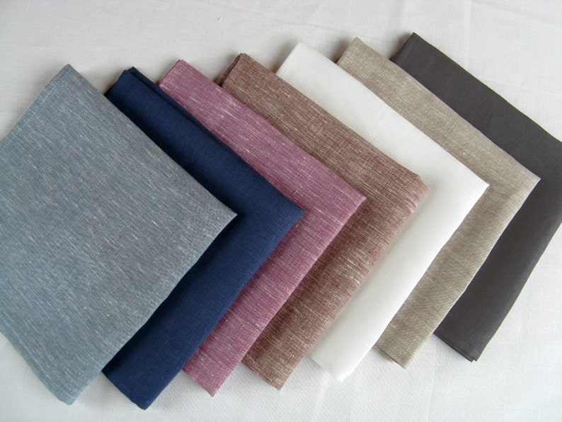 Op de afbeelding: Een set van zeven linnen servetten in verschillende kleuren, waaronder blauw, marineblauw, roze, bruin, wit, beige en grijs. De servetten zijn gevouwen en in een waaier vorm gerangschikt.