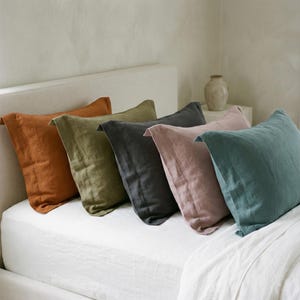 May include: Five rectangular pillowcases arranged in a row on a bed. The colors from left to right are burnt orange, olive green, charcoal gray, dusty rose, and muted teal. Each pillowcase features a visible stitched border detail along the edge.
