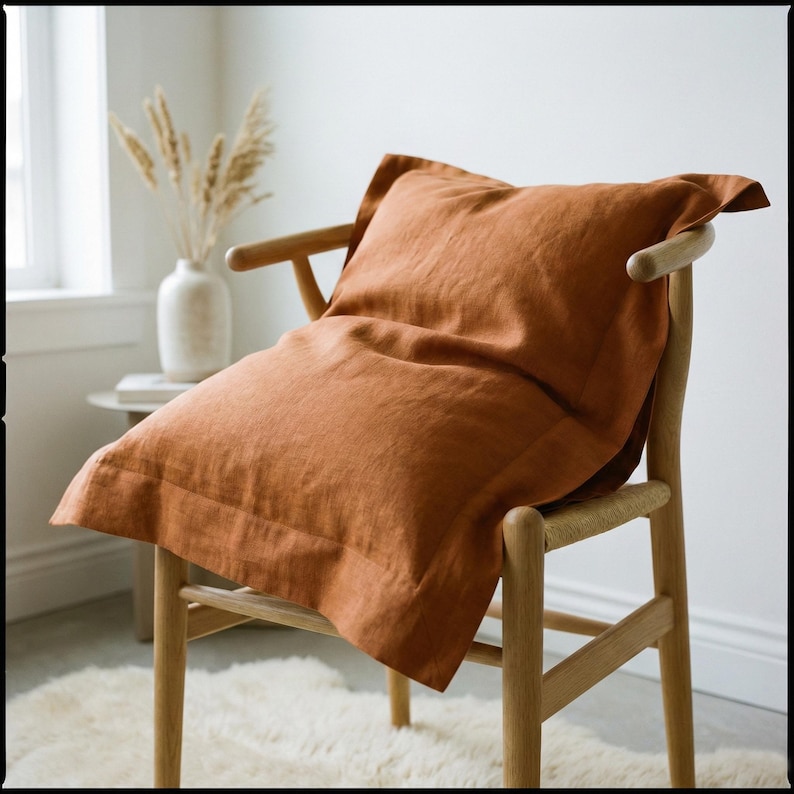 May include: A square pillowcase in a solid burnt orange color with a wide flange border. The pillowcase is draped over a light-toned wooden chair with curved armrests and a rounded backrest, showcasing a textured fabric finish throughout.