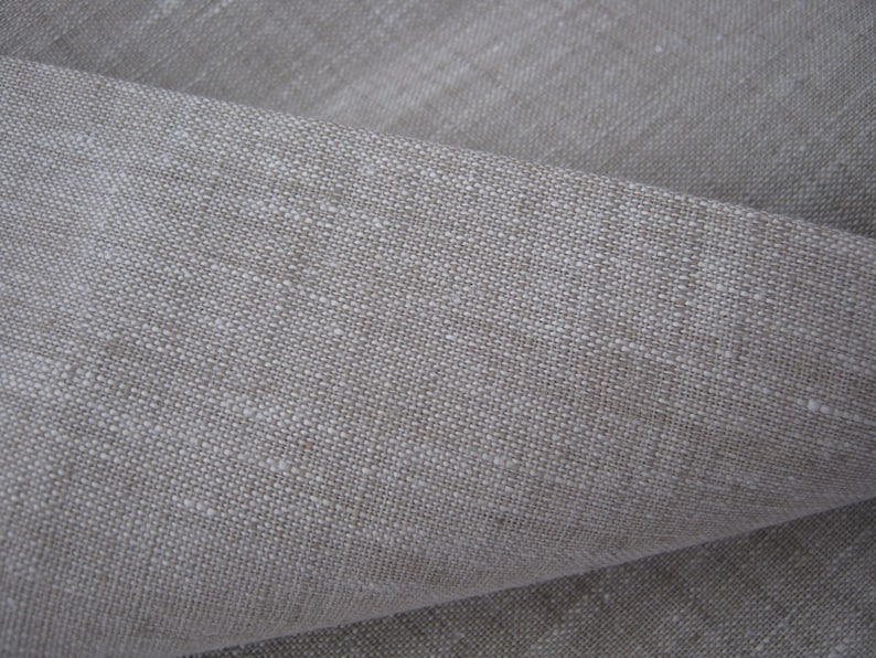 K&ouml;nnte beinhalten: Nahaufnahme eines gefalteten Abschnitts aus nat&uuml;rlichem Leinenstoff. Der Stoff hat eine neutrale, hellbeige Farbe mit einem leicht strukturierten Gewebe. Die Fasern sind sichtbar und erzeugen ein subtiles, organisches Muster. Ideal f&uuml;r Heimtextilien oder Kleidung.