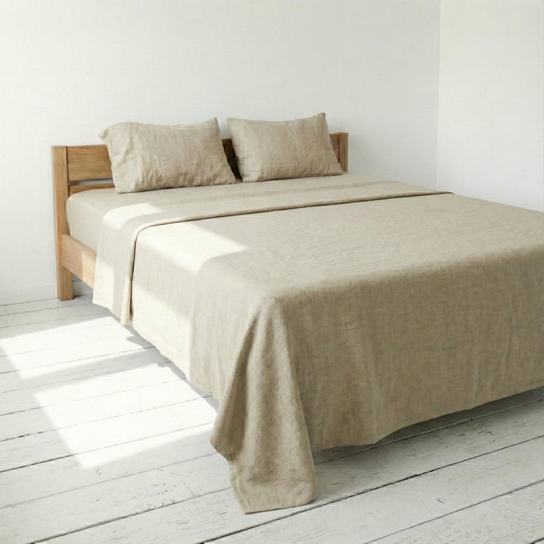 Bed with oatmeal linen sheets and pillowcases in a sunlit room with white floorboards