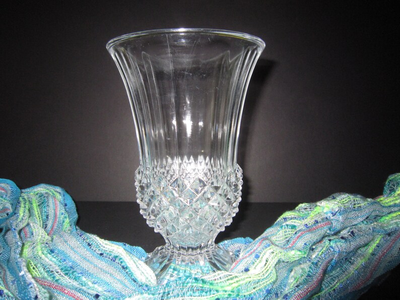 May include: A clear glass vase with a diamond pattern. The vase has a wide mouth and a narrow base. It is sitting on a blue and green patterned fabric.