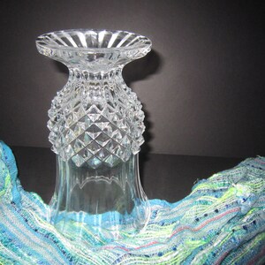 May include: A clear glass vase with a diamond pattern. The vase has a wide mouth and a narrow base. It is sitting on a blue and green patterned fabric.