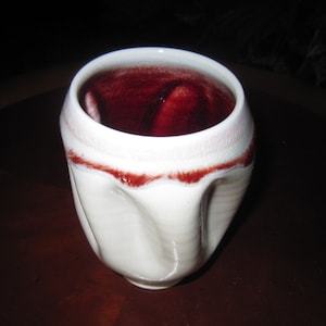 May include: White ceramic cup with a ruffled rim and a dark red glaze inside. The cup is sitting on a dark brown surface.