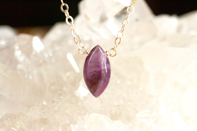May include: A delicate gold chain necklace with a purple amethyst pendant. The pendant is a marquise shape and has a natural, raw look.