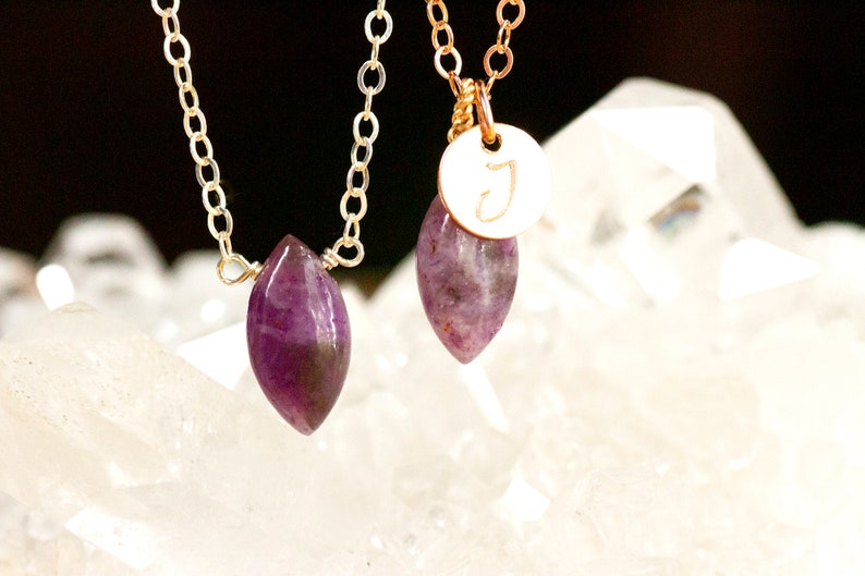 May include: Two gold necklaces with purple gemstone pendants. One pendant is a marquise-shaped stone, the other is a round stone with a gold initial "J" charm.