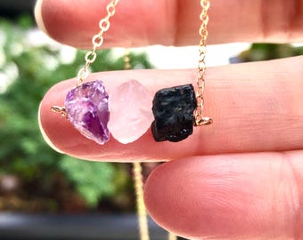 May include: A gold chain necklace with three raw crystal stones: amethyst, rose quartz, and black tourmaline.