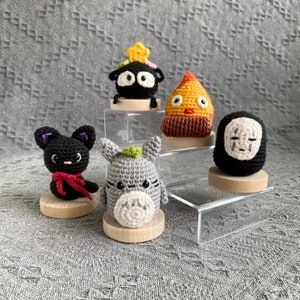 May include: A collection of handmade crochet figures, including a black cat with a red scarf, a gray Totoro-like creature, and other whimsical characters. Each figure is displayed on a small wooden base, showcasing the detailed craftsmanship and vibrant colors.