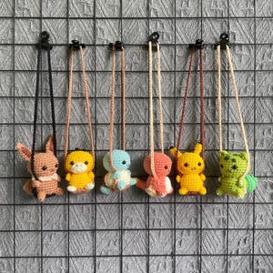 May include: A collection of seven crocheted character necklaces. Each necklace features a different character in various colors, including pink, yellow, blue, and green. The characters are suspended from a cord, and the necklaces are displayed against a gray grid background.