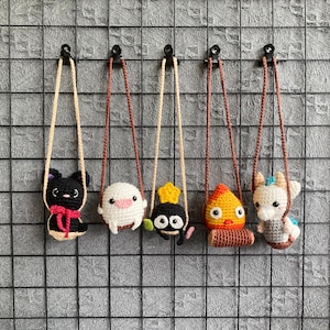 May include: Five handmade crochet necklaces featuring cartoon characters. The characters include a black cat with a red bow, a white creature, a black soot sprite with a yellow star, a yellow and red character, and a white horse with blue accents. Each necklace has a different colored cord.