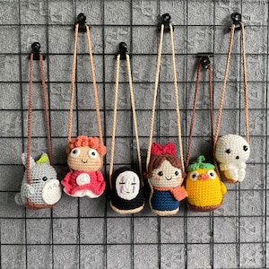 Puede incluir: Seis colgantes de personajes de ganchillo con cordones beige cuelgan contra una rejilla gris. Los personajes incluyen un Totoro gris, una figura vestida de rojo, una figura negra con una máscara blanca, una niña con un lazo rojo, un pájaro amarillo y una criatura blanca.