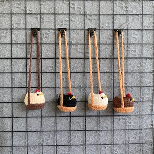 Peut inclure: Quatre ornements en forme de poulet crochetés, suspendus devant un fond quadrillé. Chaque ornement présente une combinaison de couleurs différente : marron et crème, noir, crème et beige, et marron. Ils ont des crêtes rouges et des becs jaunes.