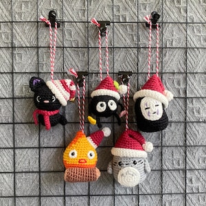May include: A collection of six crocheted ornaments featuring characters from animated films. Each ornament is wearing a red and white Santa hat and is suspended by a red and white string. The ornaments are in various colors, including black, gray, orange, and white.