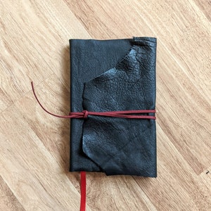 Könnte beinhalten: Ein schwarzes Lederjournal mit einem roten Lederband zum Verschließen. Das Journal ist geschlossen und liegt flach auf einer Holzoberfläche.