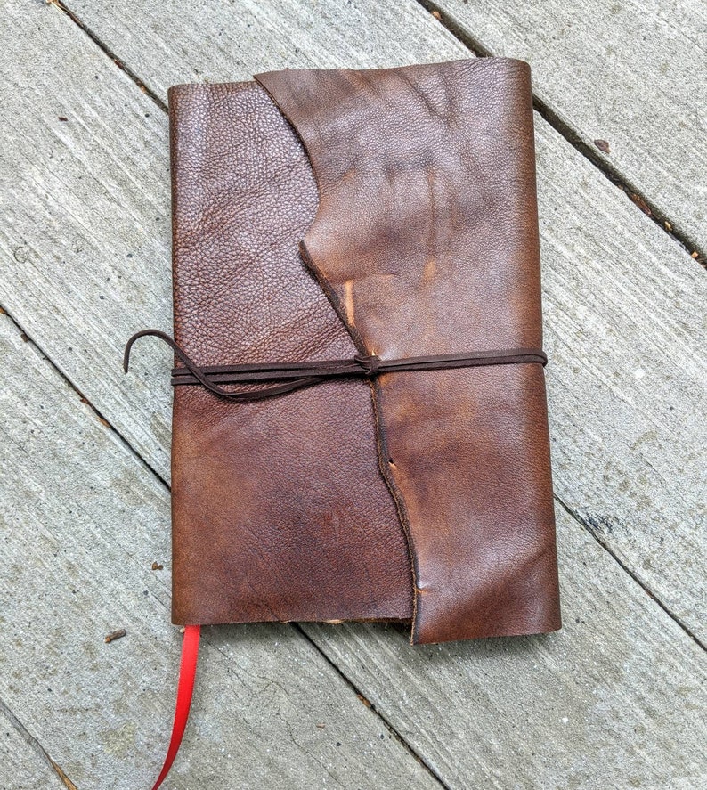 May include: A brown leather-bound journal with a red ribbon bookmark. The journal has a rustic, handmade look and is tied closed with a brown leather strap.