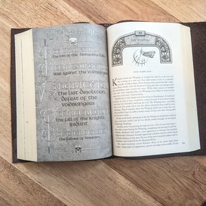 May include: A close-up of an open book with a brown leather cover. The pages are open to a section titled "The Weeping" and feature a detailed illustration of a skull and swords. The text on the page describes the "Last Desolation" and the "Fall of the Knights Radiant".