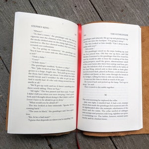 May include: Open book with a brown leather cover. The pages are open to a chapter titled "The Gunslinger" from the book "The Gunslinger" by Stephen King. The text on the page describes a scene where a gunslinger and a young boy are talking about a cellar.